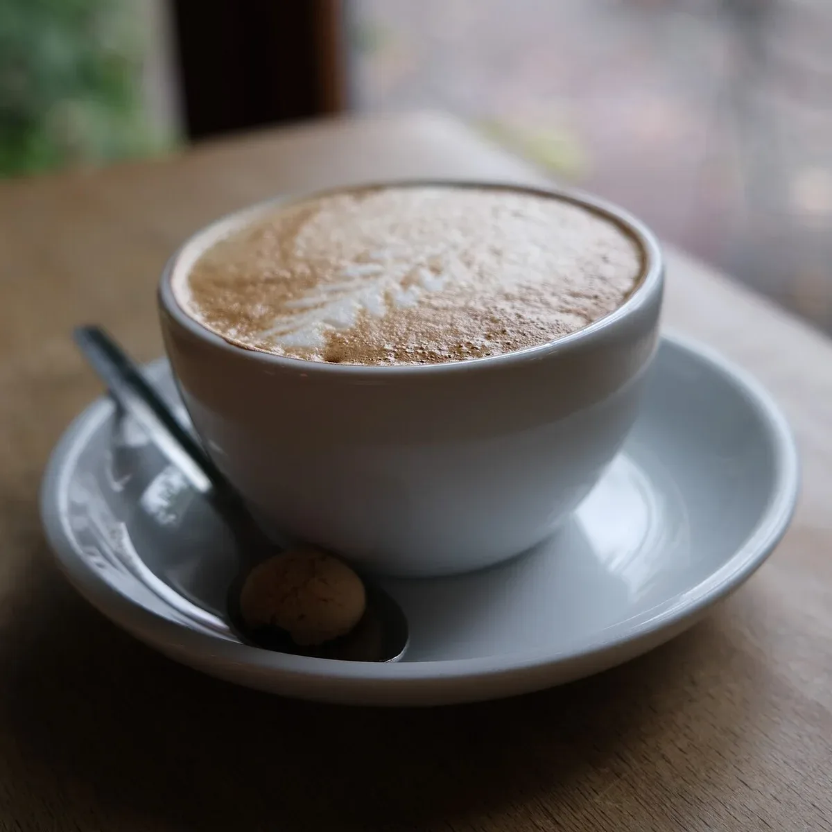 A cup of frothy coffee on a saucer beside a window