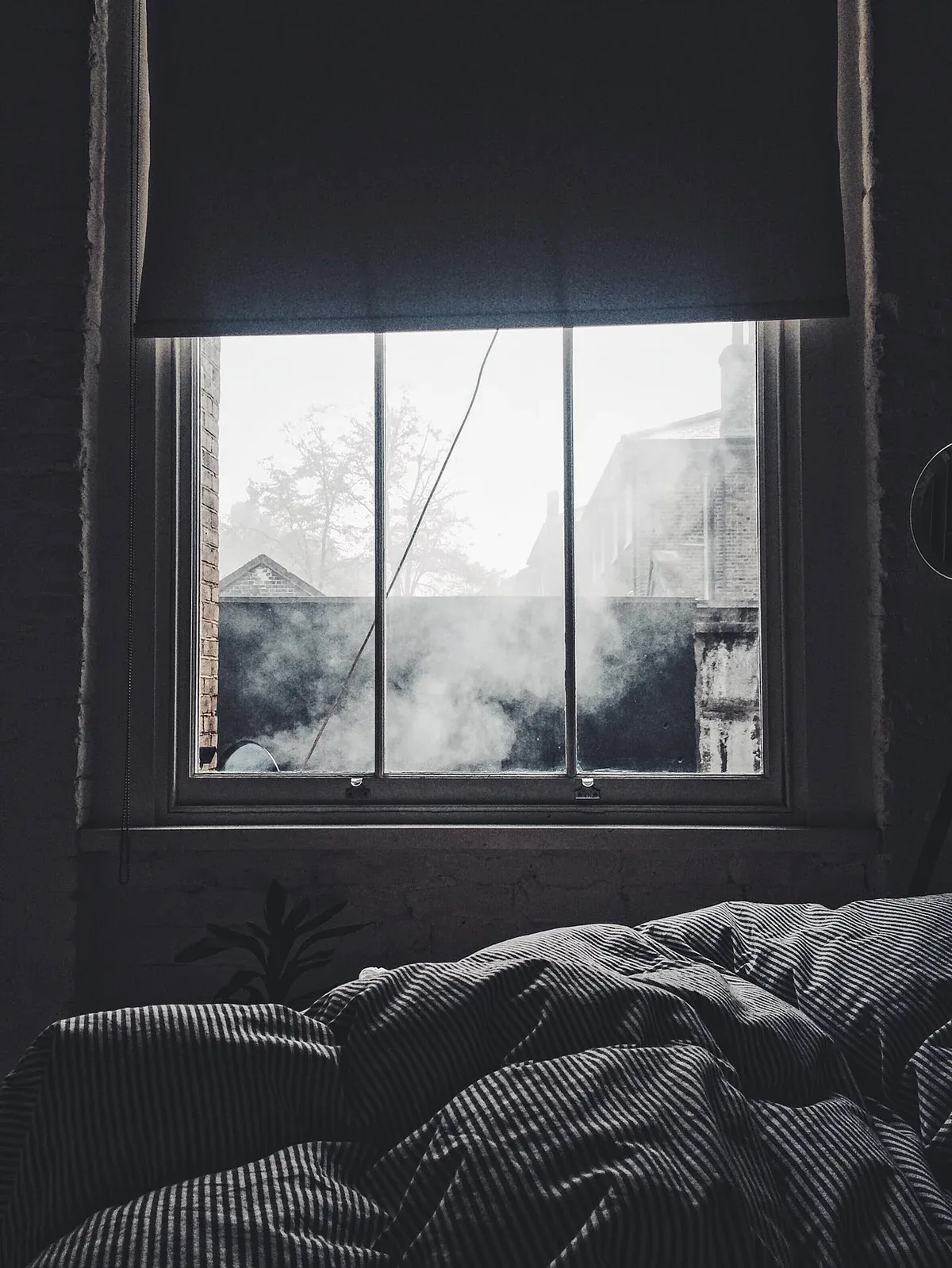 A dim bedroom with a window, lowered blind and rumpled bedding